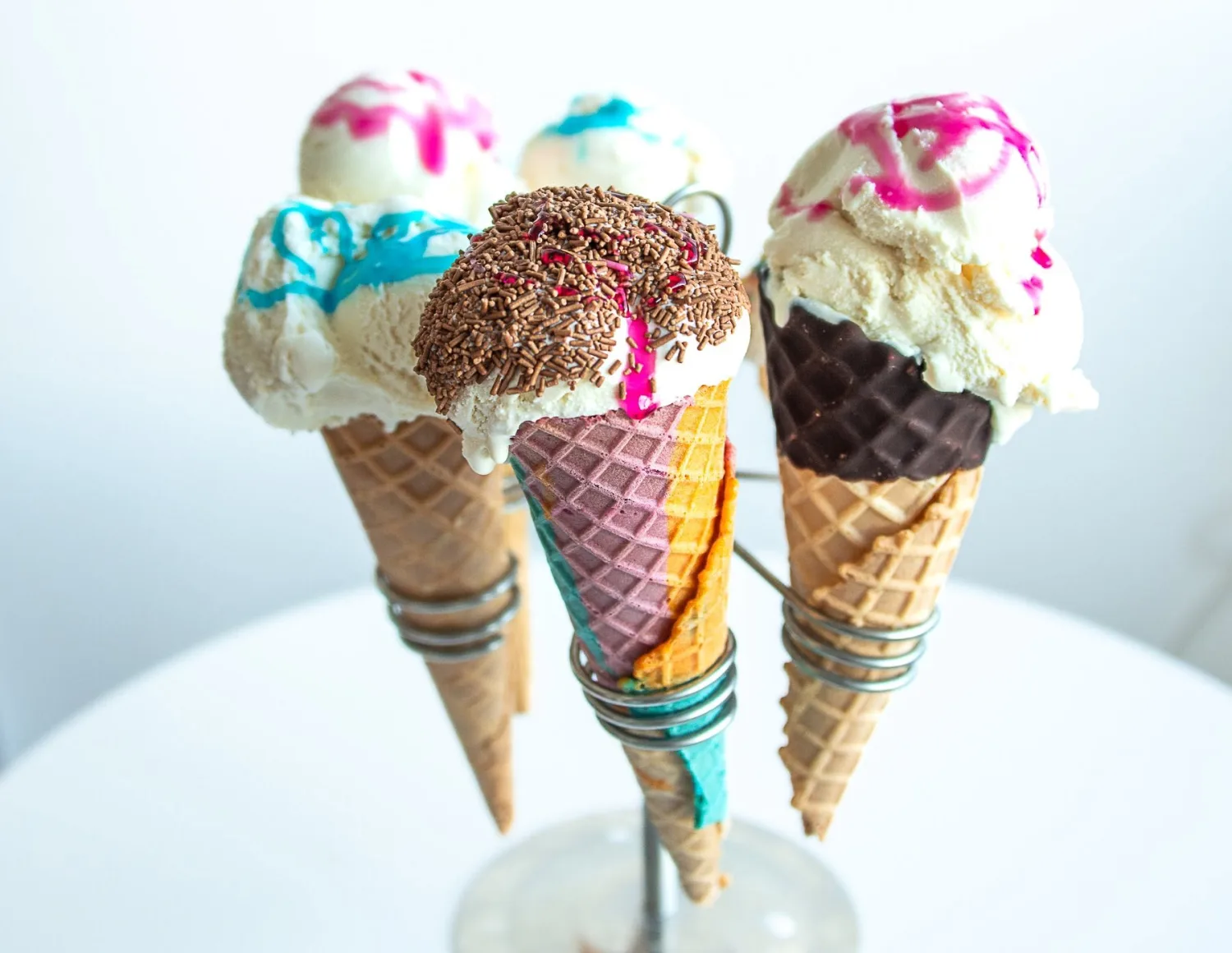 Three Manfredi vanilla ice cream cones topped with pink and blue sauce and rainbow sprinkles