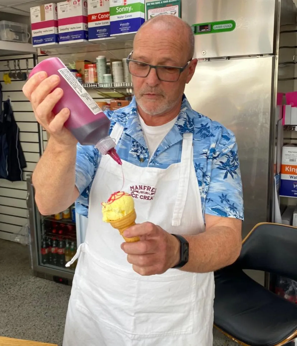 Austin Manfredi in a Hawaiian shirt and apron, drizzling sauce over a fresh cone at the shop